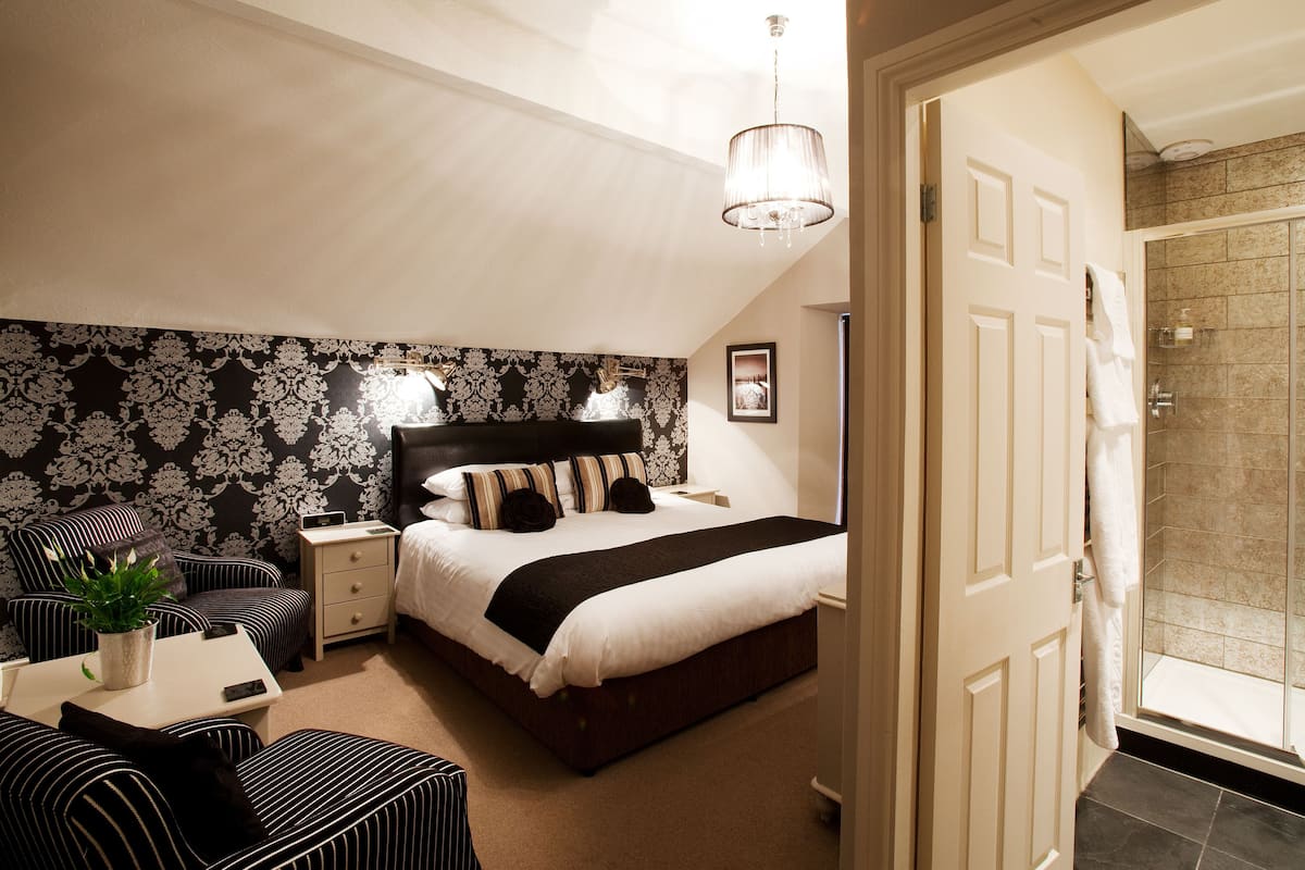A bedroom with a large bed, a bedside table, a nightstand, a chair, a potted plant, and a shower area.