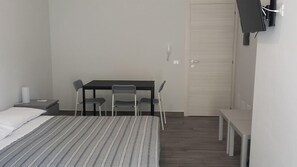 Ein kleiner, ordentlich eingerichteter Raum mit einem Bett, einem Schreibtisch, zwei Stühlen, einem an der Wand befestigten Fernseher und einer Tür.
