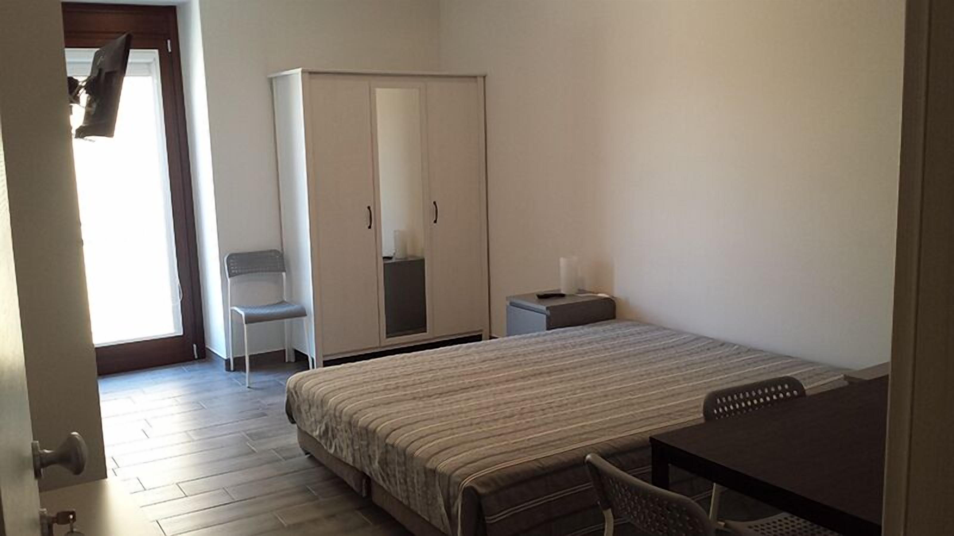 A bedroom with a bed, a desk, a chair, a wardrobe, and a small table.