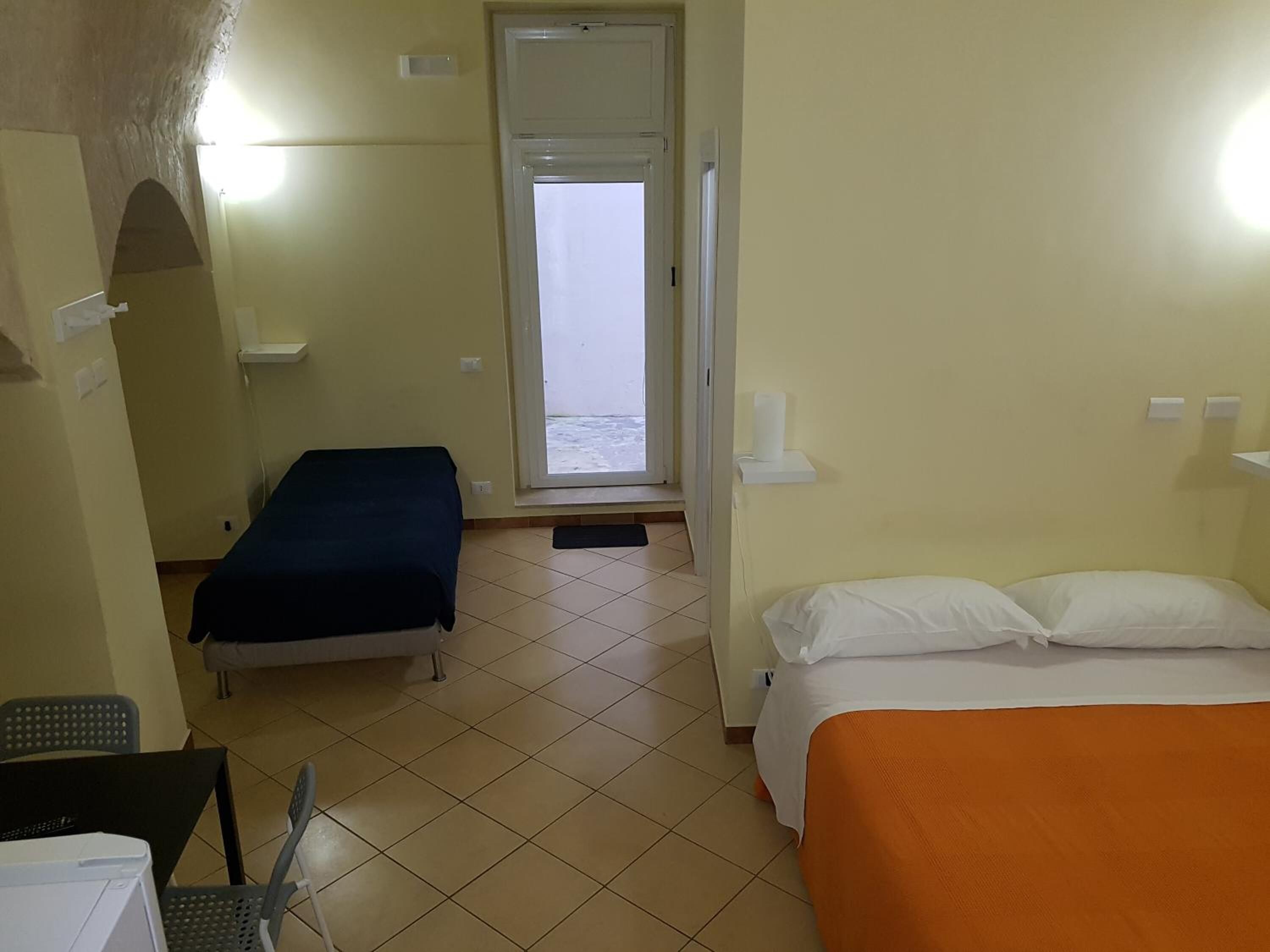 A hotel room with two beds, a window with a view of the sea, a desk with a chair, and a small table with a lamp.