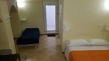 A hotel room with two beds, a window with a view of the sea, a desk with a chair, and a small table with a lamp.
