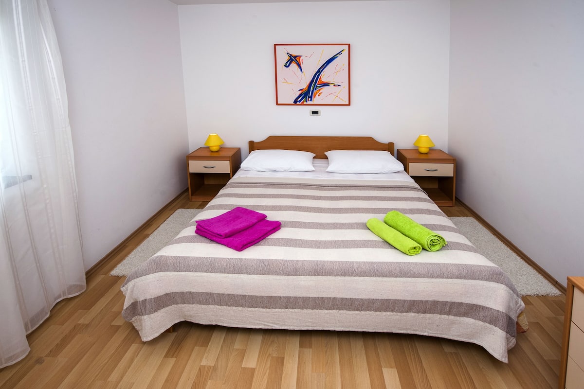 Una cama bien hecha con ropa de cama a rayas y dos almohadas, dos mesitas de noche con lámparas, una obra de arte abstracta enmarcada en la pared y un suelo de madera.