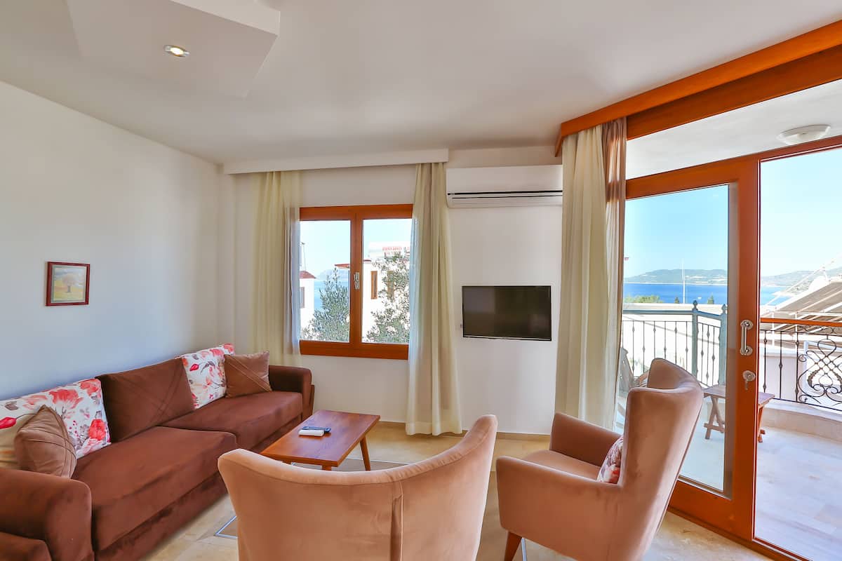 A living room with a sofa, two armchairs, a coffee table, and a TV. There is a balcony with a view of the sea.