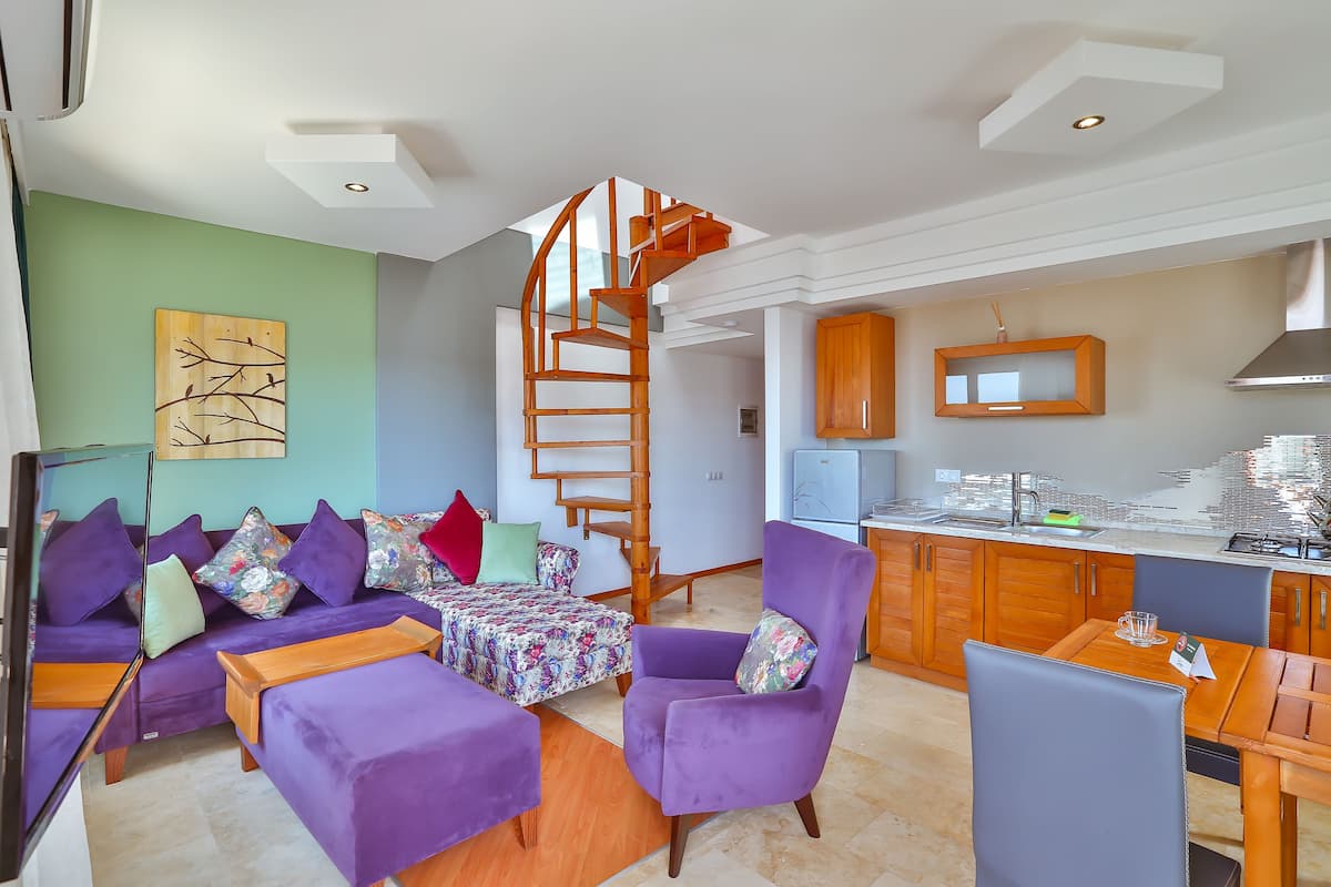 A modern living area with a sofa set, a purple armchair, a wooden dining table, and a kitchen with wooden cabinets and a spiral staircase.