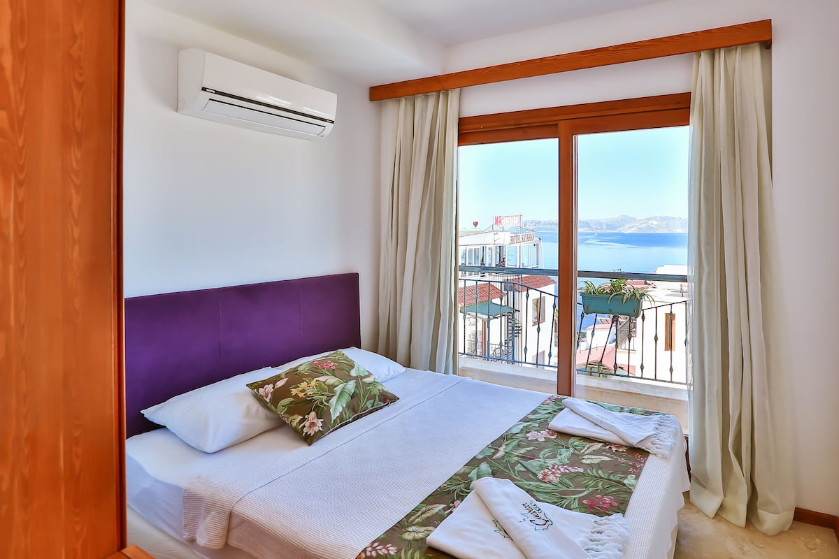 A hotel room with a bed, a window with curtains, an air conditioning unit, and a view of the sea and buildings.