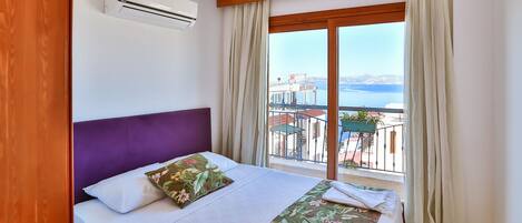 A hotel room with a bed, a window with curtains, an air conditioning unit, and a view of the sea and buildings.