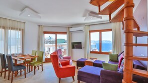 A spacious living area with a wooden staircase, a dining table with chairs, a sofa set, and large windows offering a view of the sea. - Cinarlar Apart Hotel (Kas)