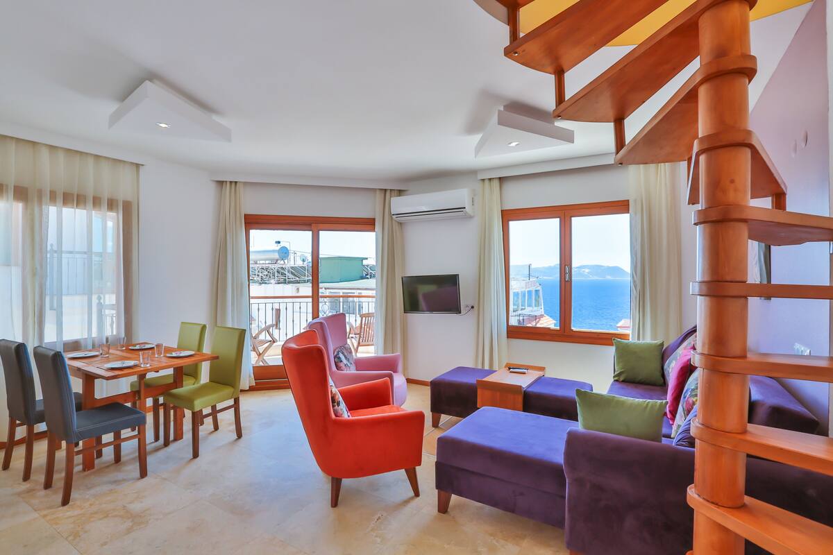 A spacious living area with a wooden staircase, a dining table with chairs, a sofa set, and large windows offering a view of the sea.