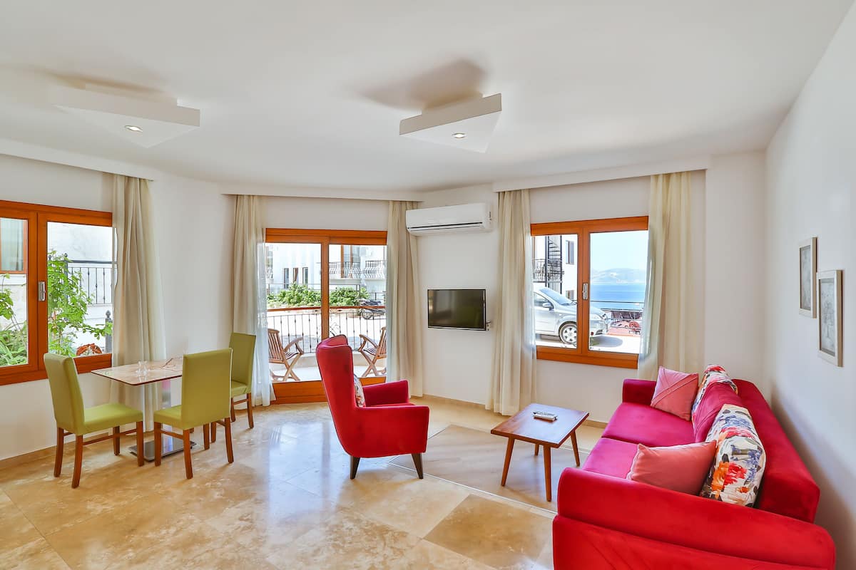 A spacious living room with a red sofa, green chairs, a small table, and large windows offering a view of the outdoors.