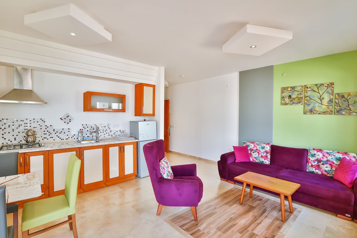A modern kitchen with white cabinets, a built-in oven, and a stainless steel range hood. There is a dining area with a wooden table and chairs, and a purple sofa with floral cushions.
