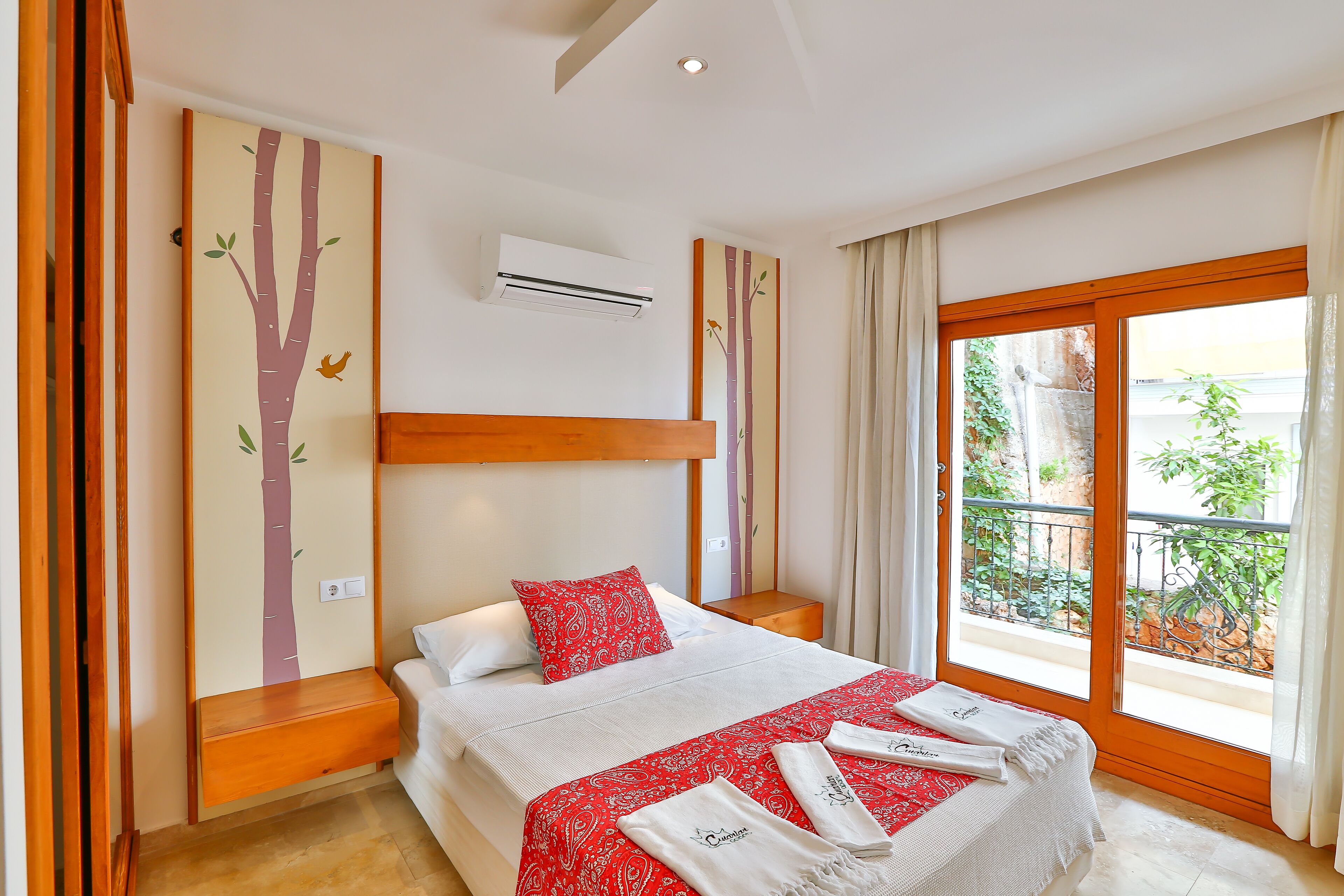 A hotel room with a large bed, a wooden headboard, a window with a view of greenery, and a wall-mounted air conditioner.
