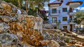 A stone water feature with a small stream, surrounded by large rocks and vegetation, in front of a multi-story building with balconies and a red roof. - Cinarlar Apart Hotel (Kas)