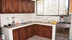 A kitchen with wooden cabinets, a stainless steel stove, and a window with sheer curtains. - Hotel Residencial El Viajero (Tababela)