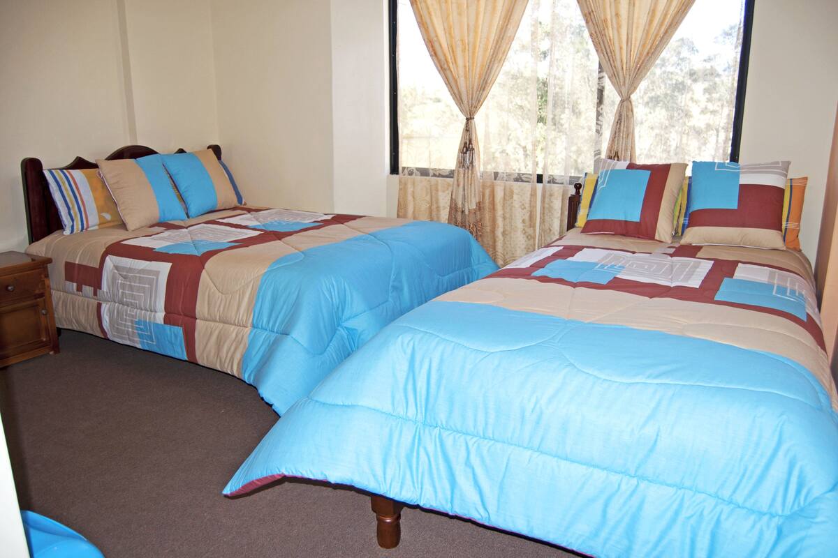 Una cama doble con colcha azul y beige, una mesita de noche y una ventana con cortinas.