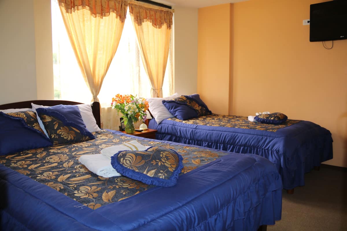 Habitación de hotel con dos camas, cada una con ropa de cama azul y almohadas decorativas. Hay un televisor montado en la pared y una ventana con cortinas.