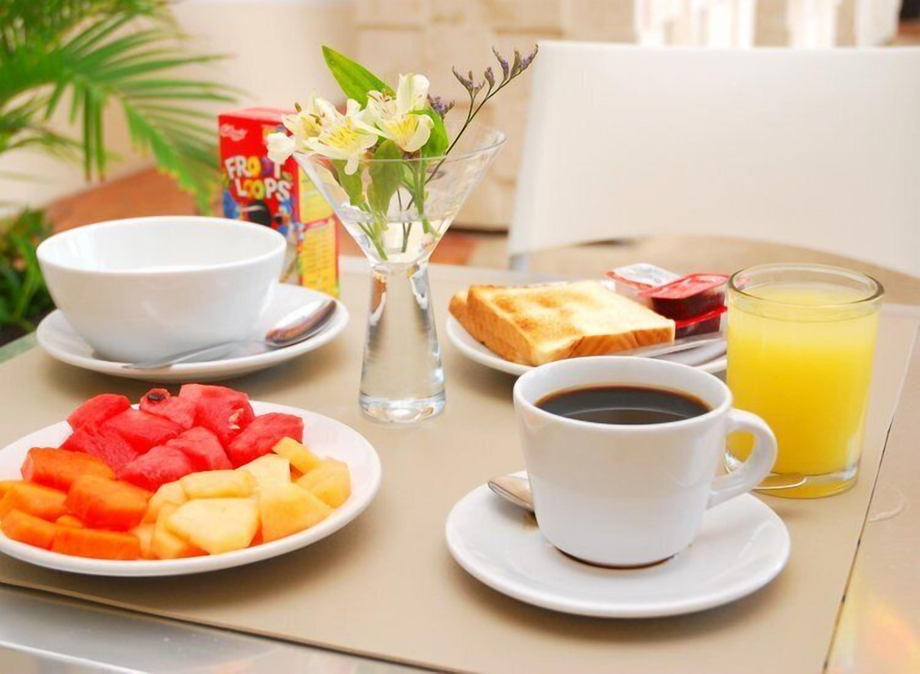 Una escena de desayuno con un plato de fruta, una taza de café, tostadas, un vaso de zumo de naranja y un florero con flores.