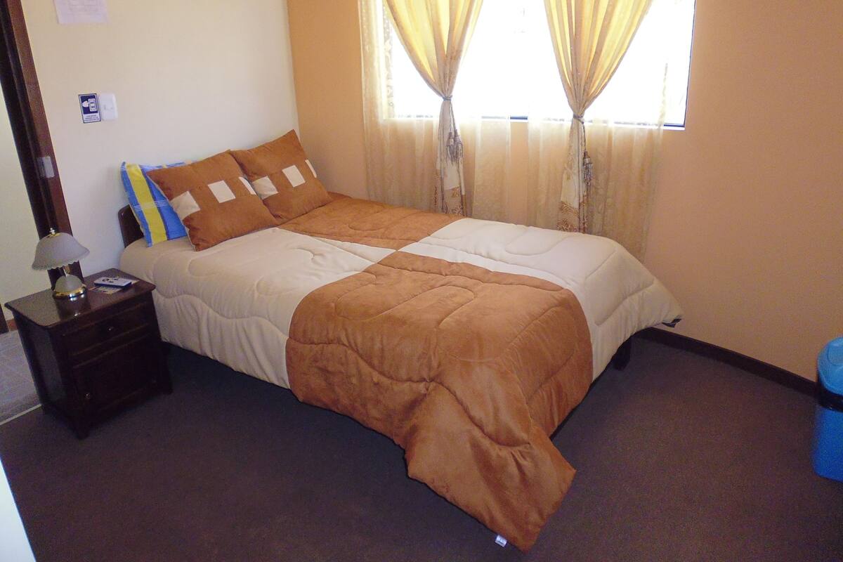 Una cama bien hecha con edredón marrón y almohadas, una mesita de noche con lámpara y una ventana con cortinas.