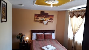 A neatly made bed with a patterned bedspread, a bedside lamp, and a painting of a sunset on the wall. - Hotel Residencial El Viajero (Tababela)