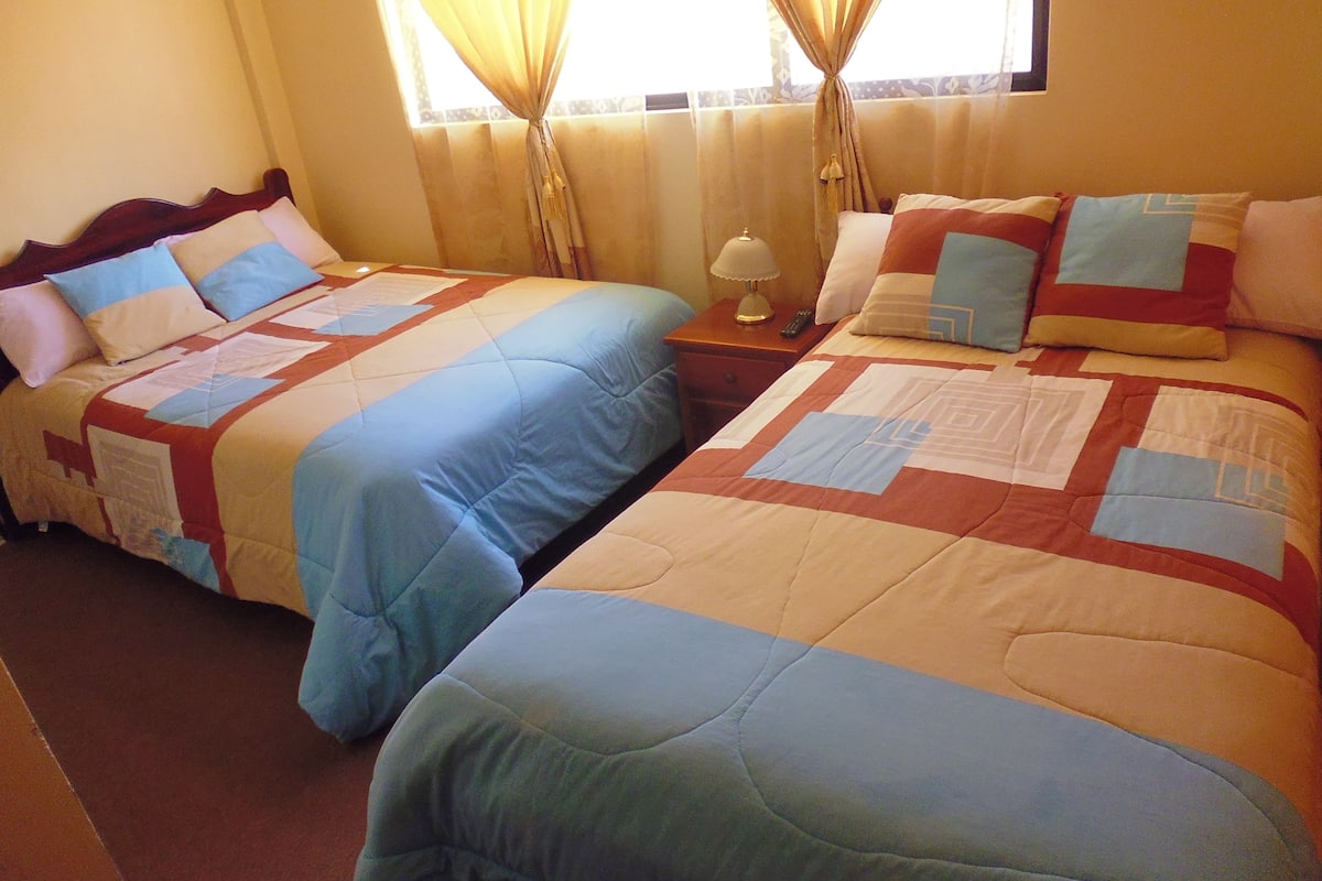 Habitación de hotel con dos camas, cada una con cubrecama acolchado y almohadas. Hay una mesita de noche con una lámpara y un control remoto.