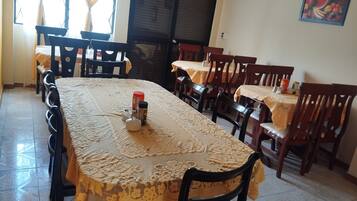 Un comedor con una mesa larga cubierta con un mantel de encaje, rodeada de sillas de madera. Hay condimentos sobre la mesa y un cartel con la palabra 'SALIDA' en la pared.