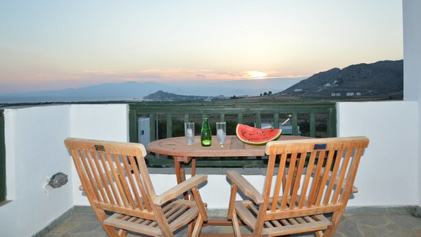 Ein Holztisch mit zwei Stühlen, einem Glas Wasser, einer Flasche und einem Melonenscheibchen auf einem Balkon mit Sonnenuntergang und Blick auf ferne Berge.