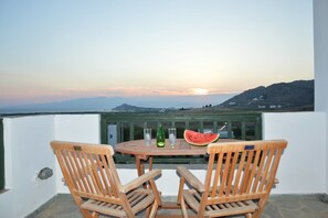 Ein Holztisch mit zwei Stühlen, einem Glas Wasser, einer Flasche und einem Melonenscheibchen auf einem Balkon mit Sonnenuntergang und Blick auf ferne Berge.