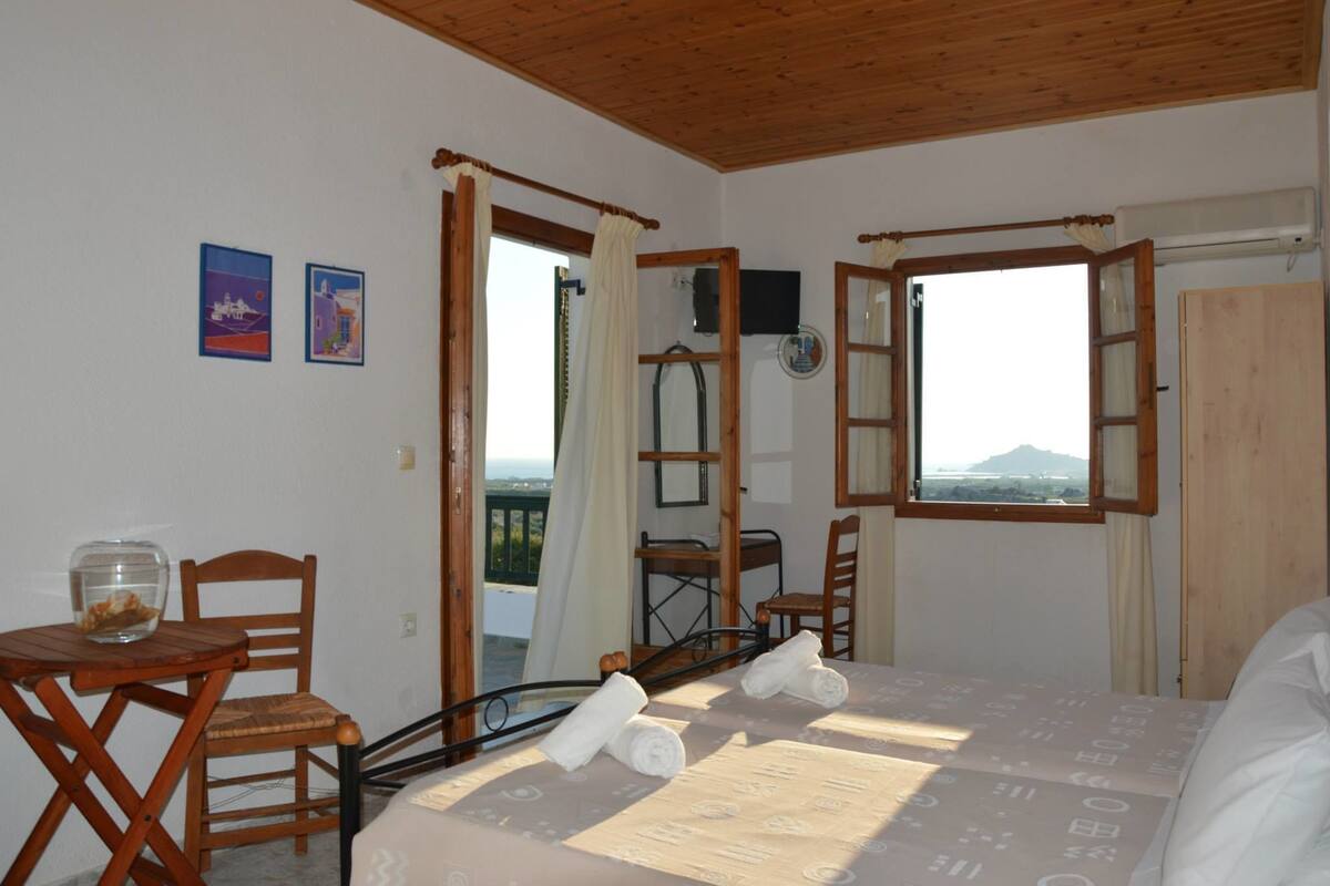 A room with a bed, two chairs, a table with a fishbowl, and a view of the sea.