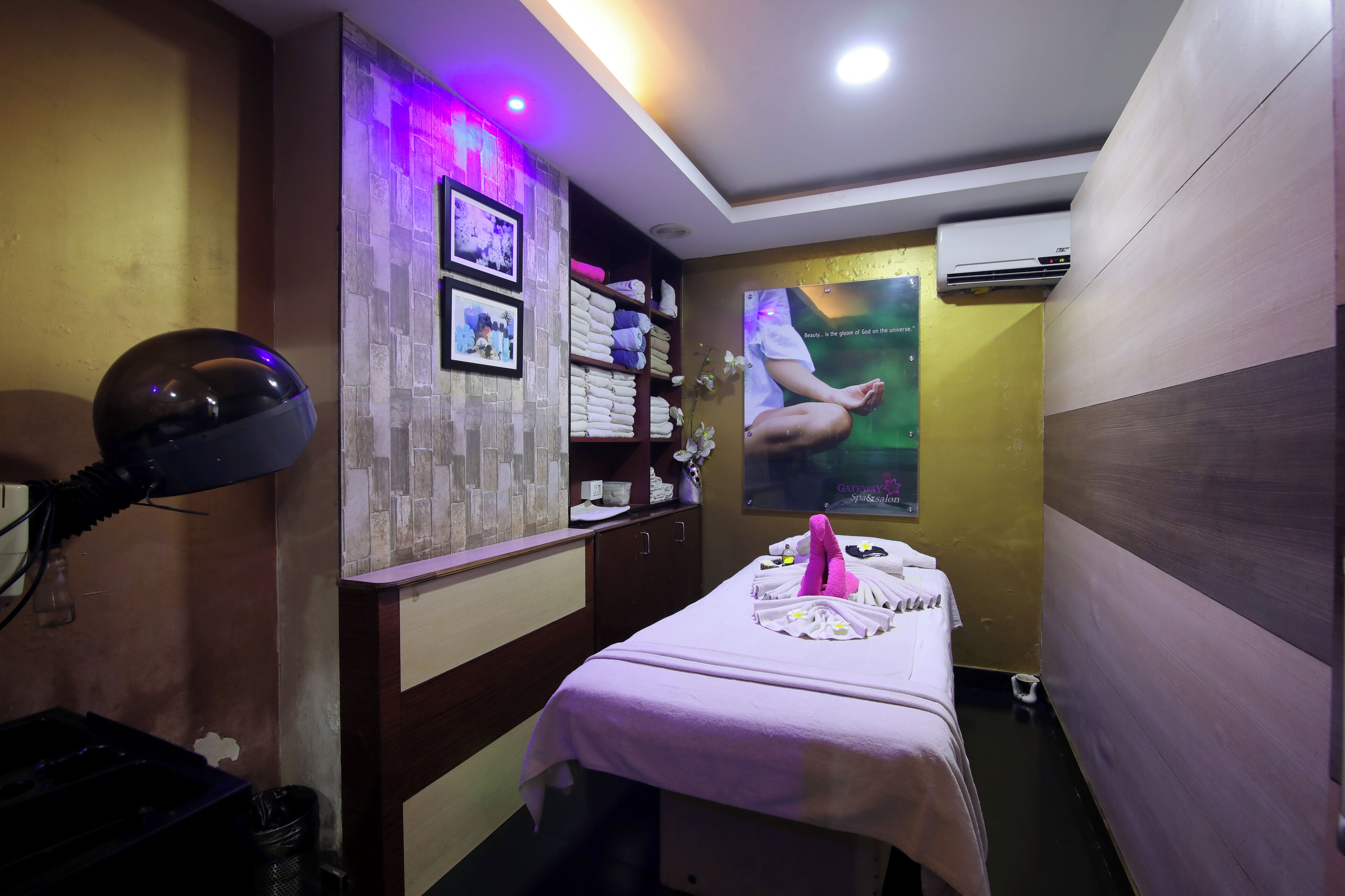 Ein Massageroom mit Massagetisch, einem an der Wand befestigten Massagesessel und einem Regal mit Handtüchern und einer kleinen Pflanze.
