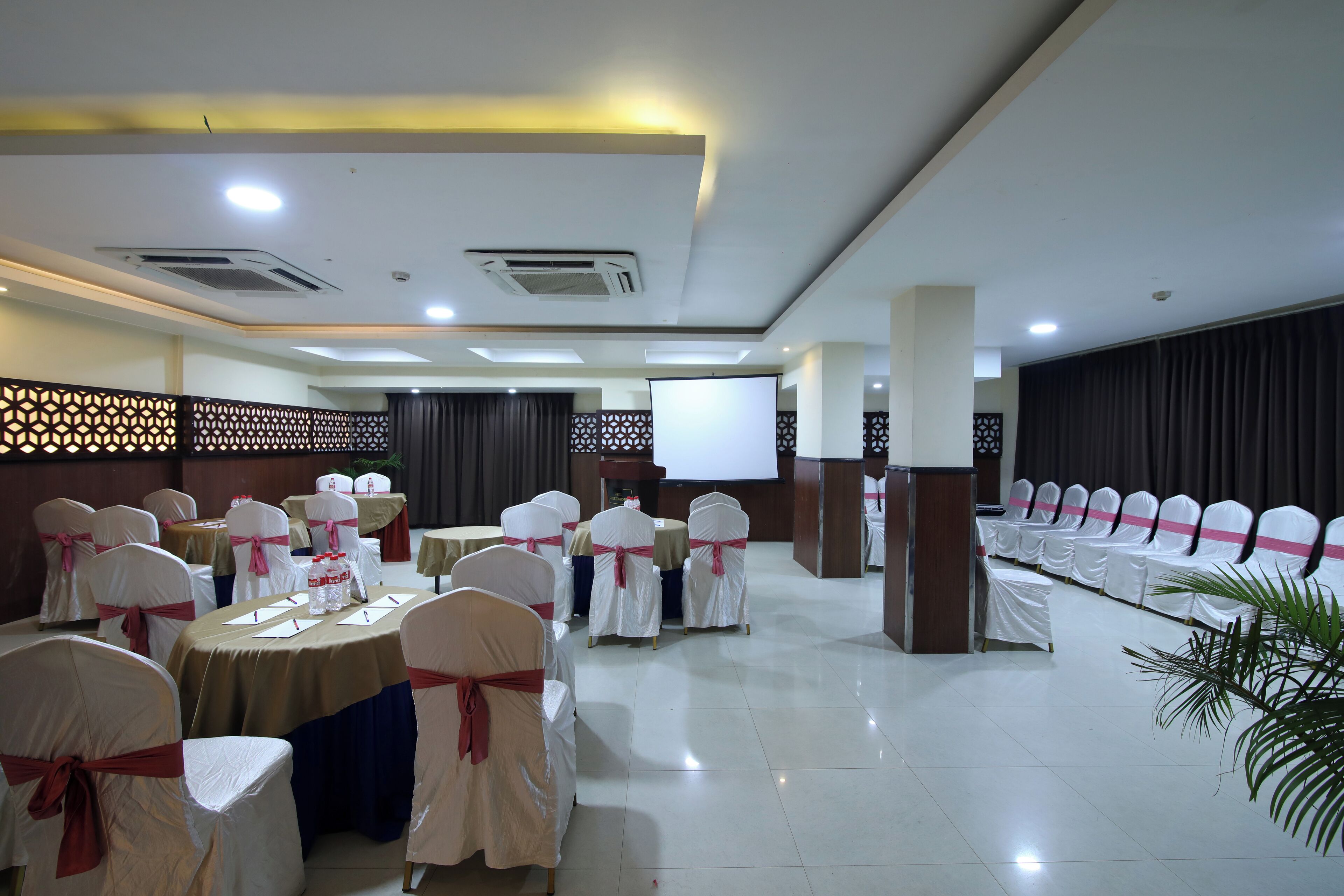 A conference room with rows of white chairs, tables with brown tablecloths, and a large screen at the front.