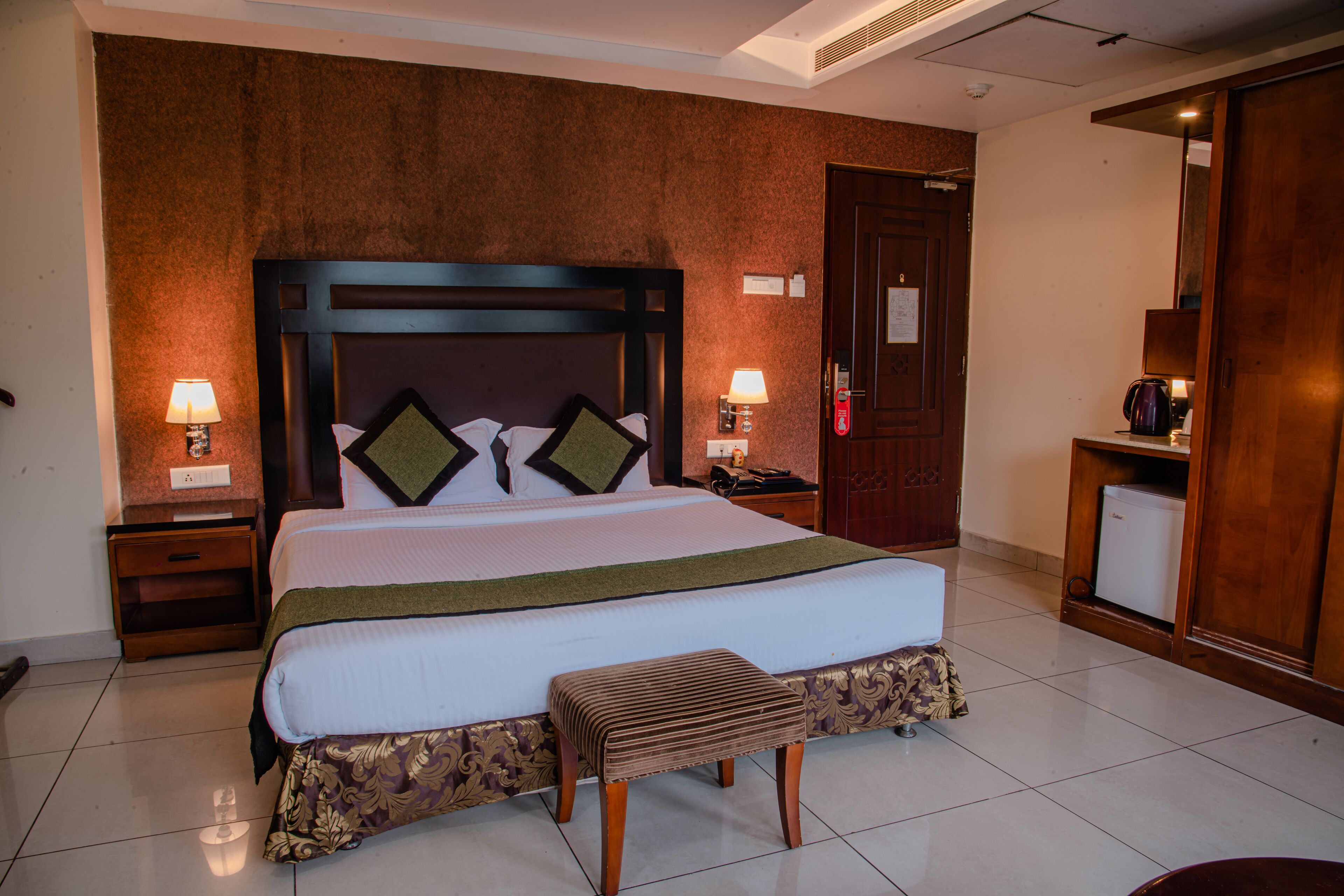 Une chambre d’hôtel avec un grand lit, une tête de lit en bois, deux tables de chevet avec des lampes, une petite table avec une bouilloire et une armoire.