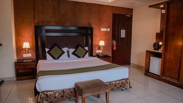 A hotel room with a large bed, wooden headboard, two bedside tables with lamps, a small table with a kettle, and a wardrobe.