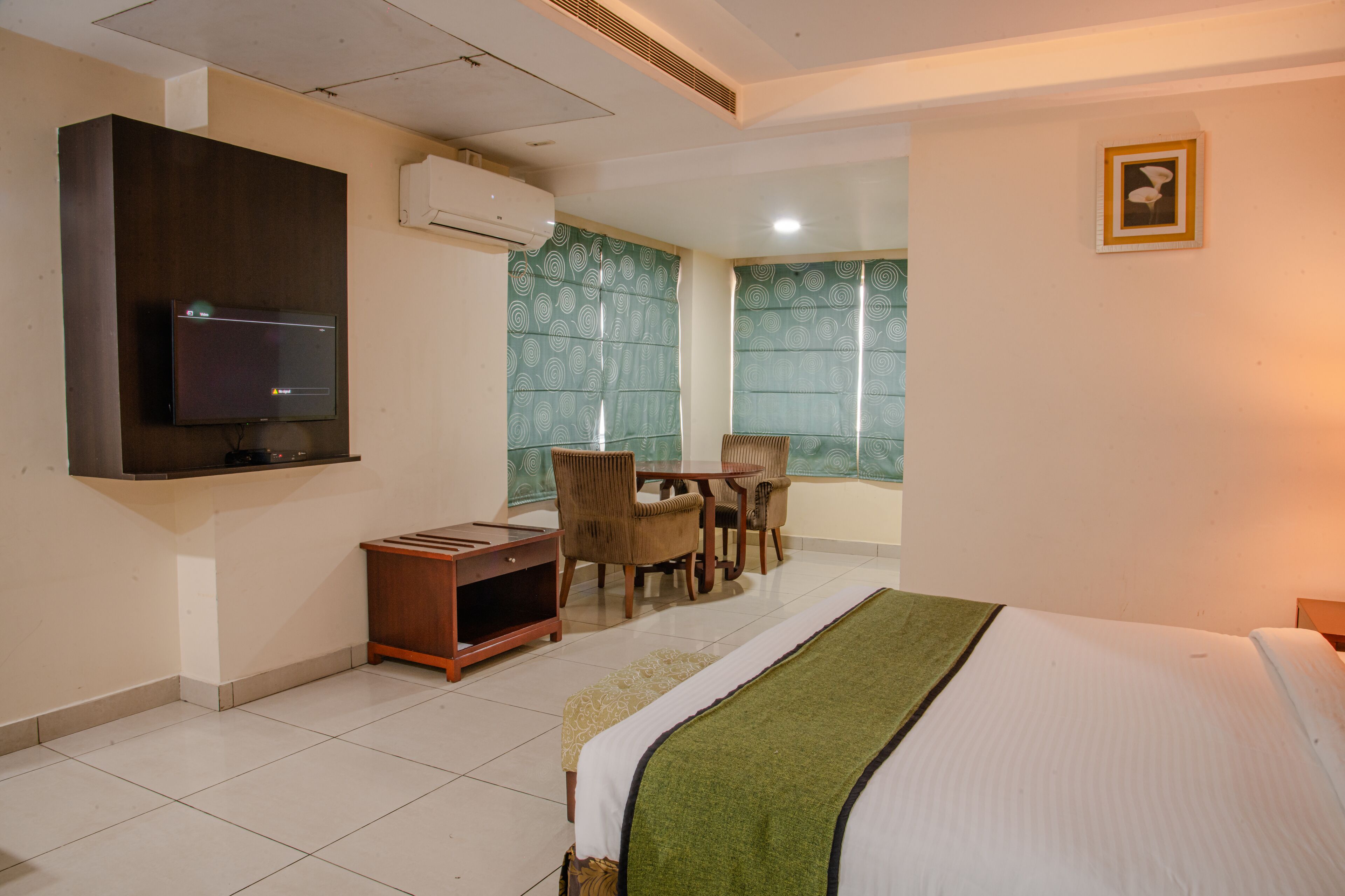 A hotel room with a bed, a TV mounted on the wall, a small desk with chairs, and a window with patterned curtains.