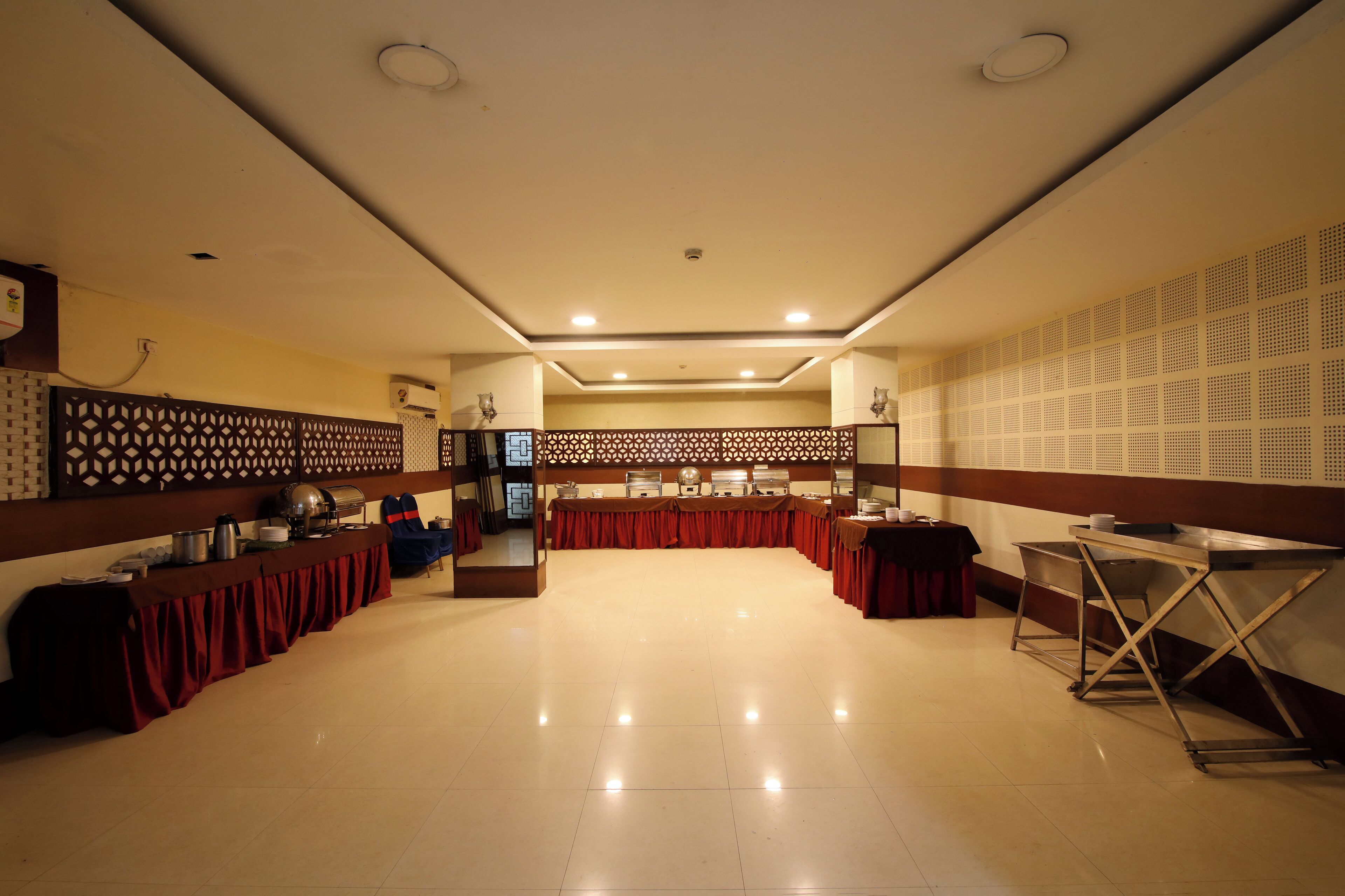 A spacious banquet hall with a long table covered in a red tablecloth, equipped with a buffet setup on one side and a service counter on the other.