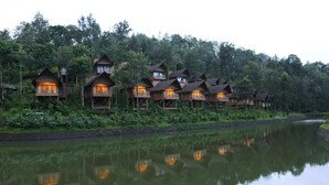 A row of wooden cabins with balconies, situated by a calm lake, surrounded by dense forest. - Kofiland Resort (Peermade)