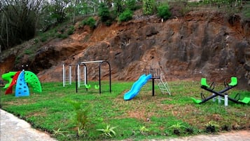 Explore the playground area with a slide, swings, and climbing structures set amidst lush greenery and a scenic hillside backdrop.