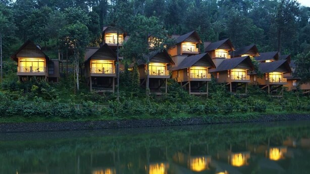 A row of wooden cabins with balconies, surrounded by dense forest and a calm lake. - Kofiland Resort (Peermade)
