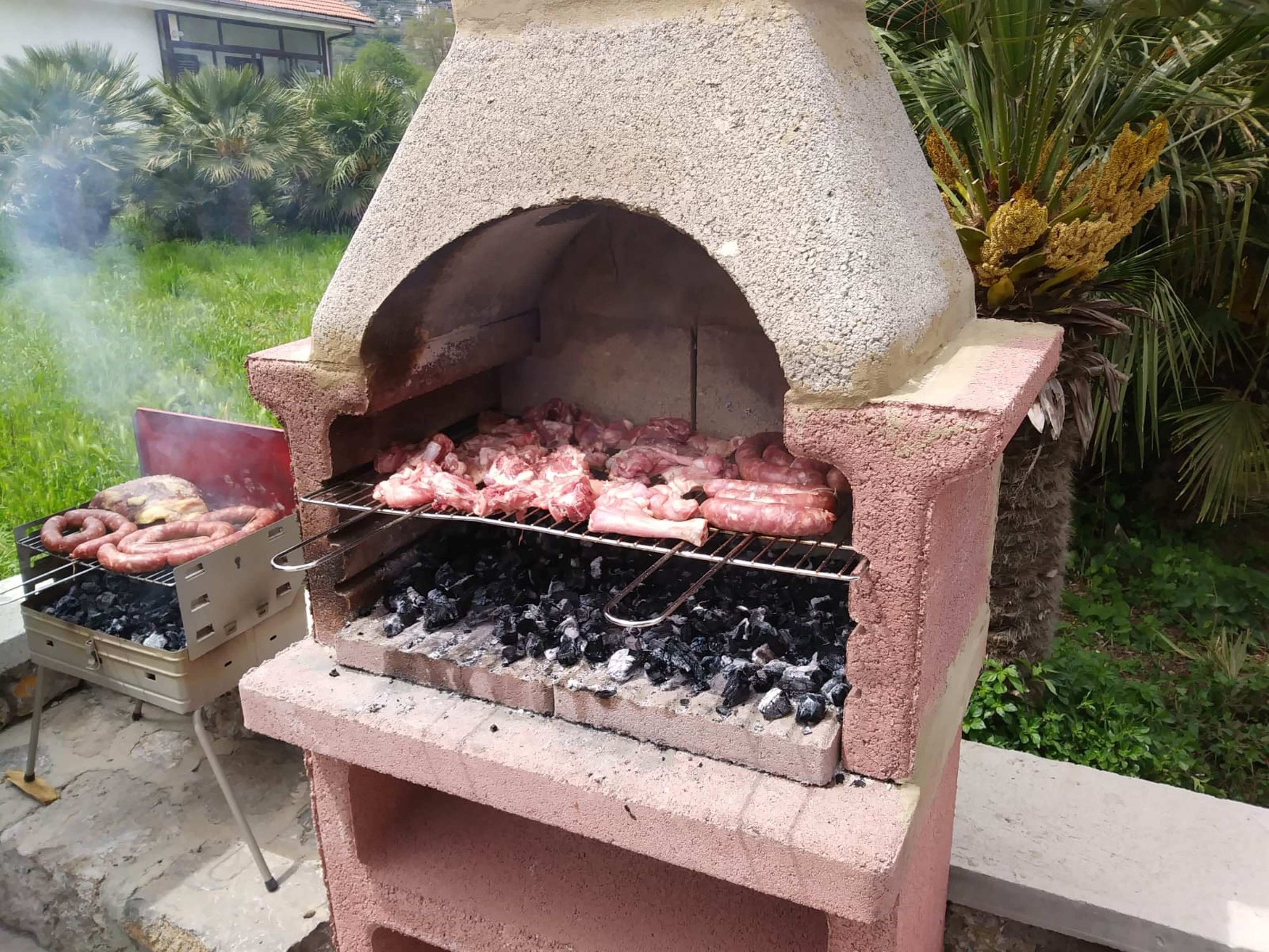 A concrete outdoor grill with a chimney, cooking sausages and chicken, and a charcoal grill with sausages.