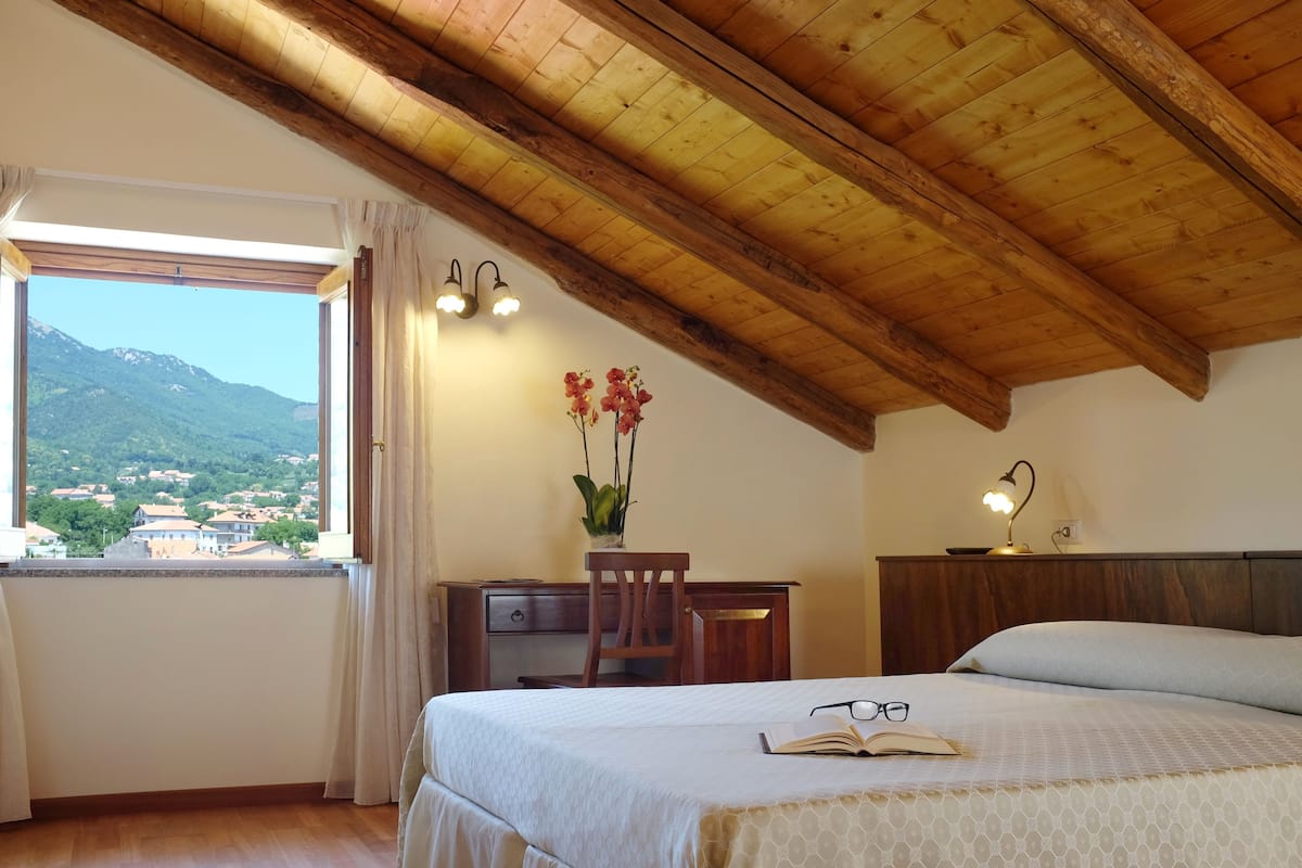 A bedroom with a wooden ceiling, a bed, a desk, and a window with a view of mountains.