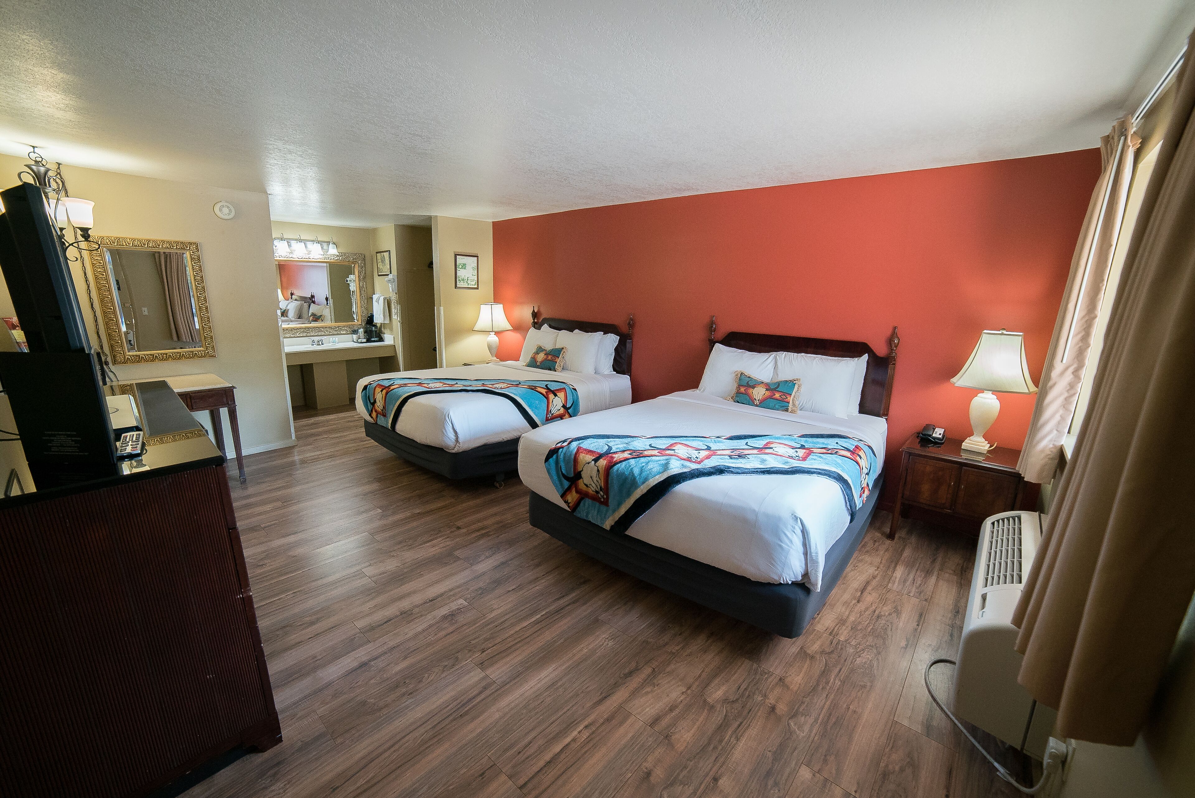 A hotel room with two beds, wooden flooring, a mirror, a desk, and a lamp.