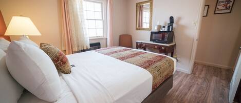 A hotel room with a large bed, a desk with a computer, a chair, a window with curtains, and a door.