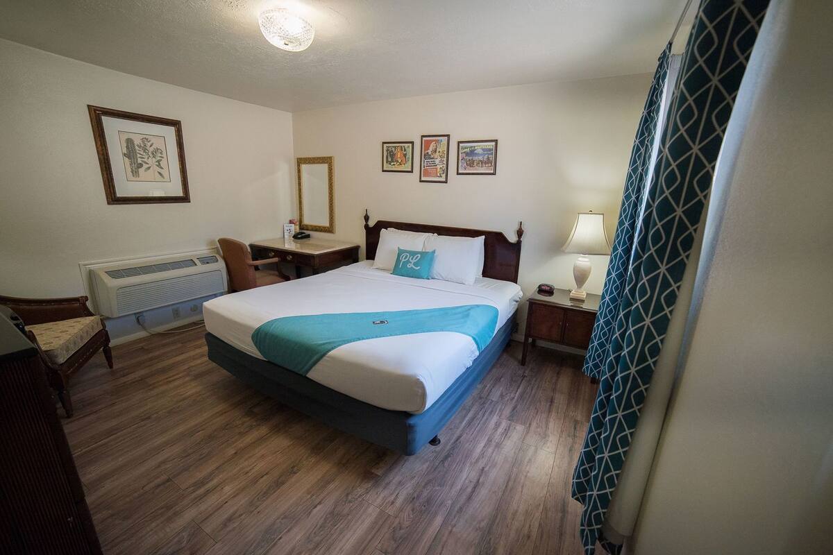 A hotel room with a wooden floor, a bed with a blue accent, a desk with a chair, a bedside table with a lamp, and a radiator.
