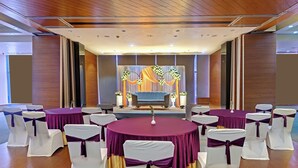 A conference room with round tables, chairs with white and purple sashes, a stage with floral decorations, and a wooden ceiling. - Hotel Suba Elite Vadodara (Vadodara)