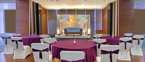 A conference room with round tables, chairs with white and purple sashes, a stage with floral decorations, and a wooden ceiling.