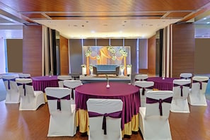 Une salle de conférence avec des tables rondes, des chaises ornées de rubans blancs et violets, une scène décorée de fleurs et un plafond en bois.