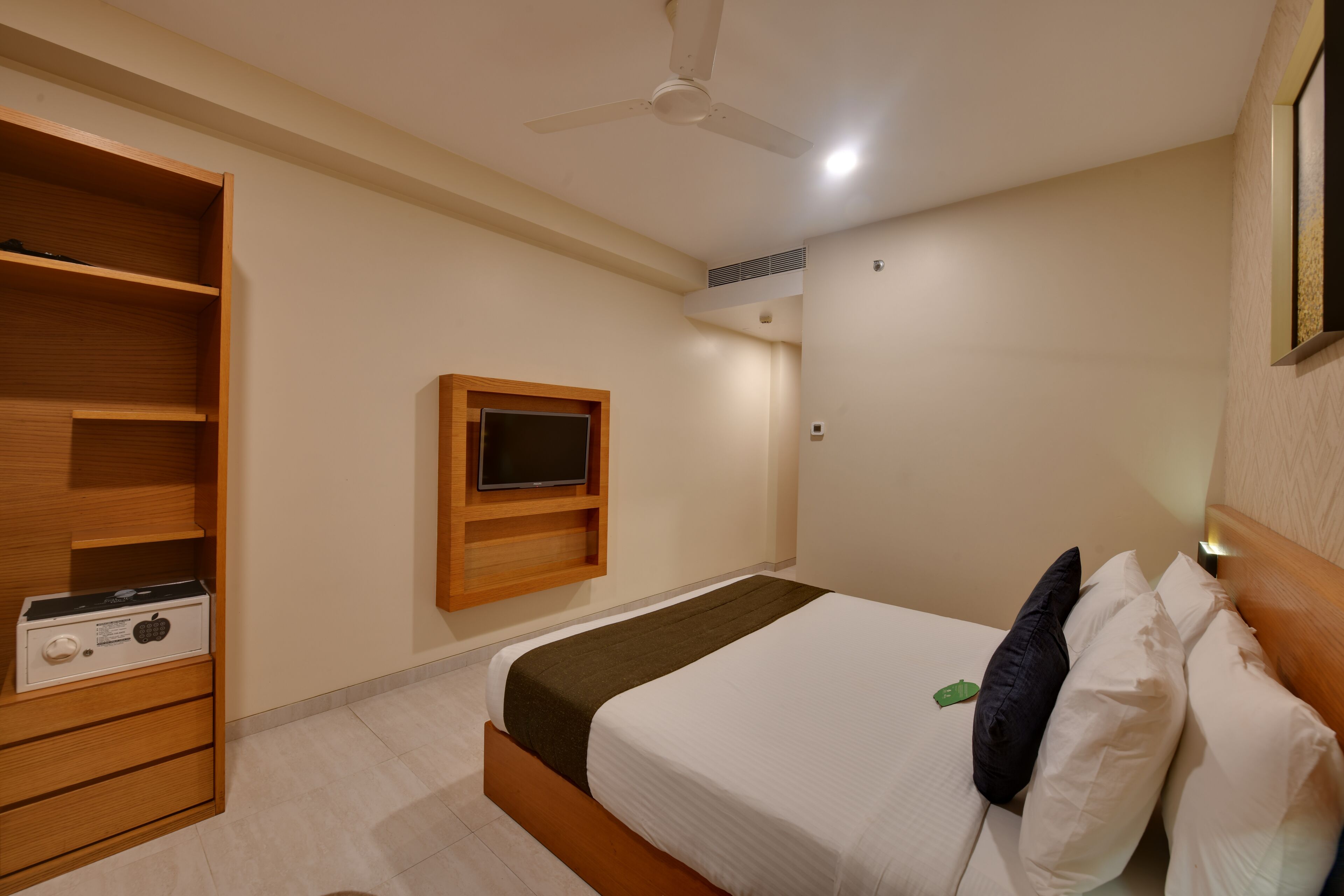 A hotel room with a bed, a TV mounted on the wall, a ceiling fan, and a wooden wardrobe.