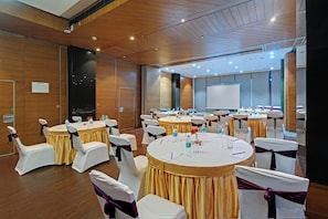 Une salle de conférence dotée de plusieurs tables rondes, de chaises blanches ornées de rubans violets et d’un grand écran de projection.