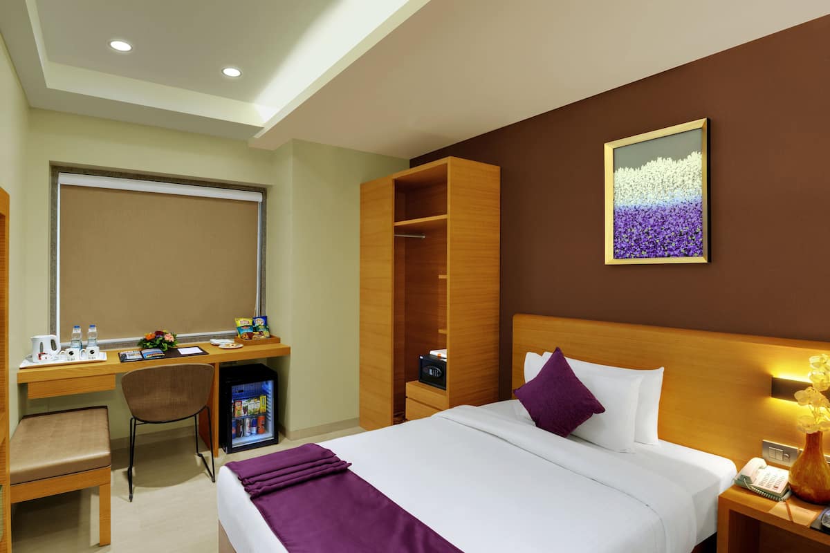 A hotel room with a bed, desk, chair, minibar, and a painting on the wall.