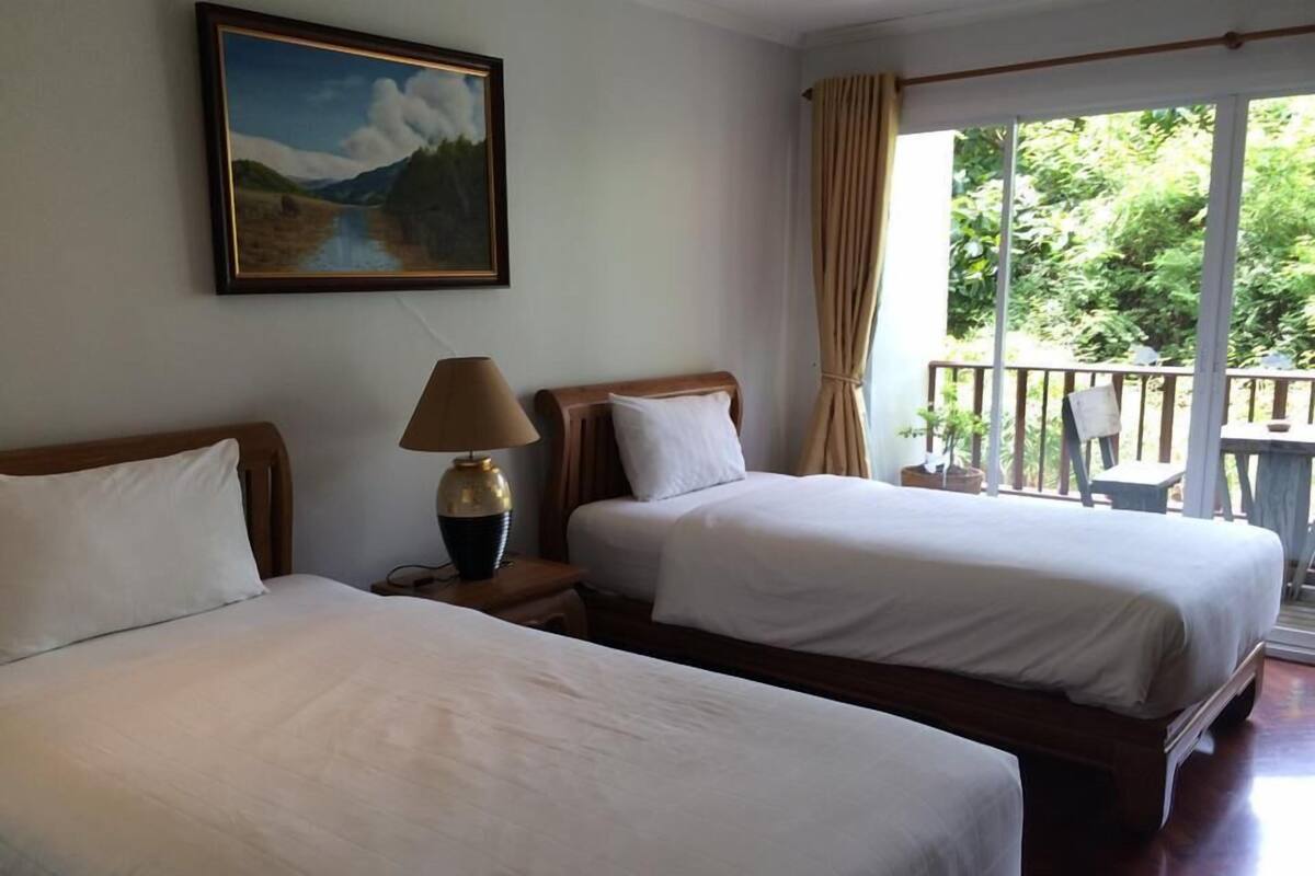 A hotel room with two beds, a painting on the wall, a lamp, and a balcony with a view of greenery.
