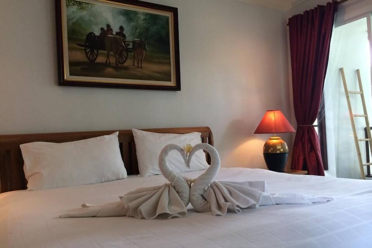 A hotel room with a neatly made bed, a painting on the wall, a lamp, and a window with curtains.