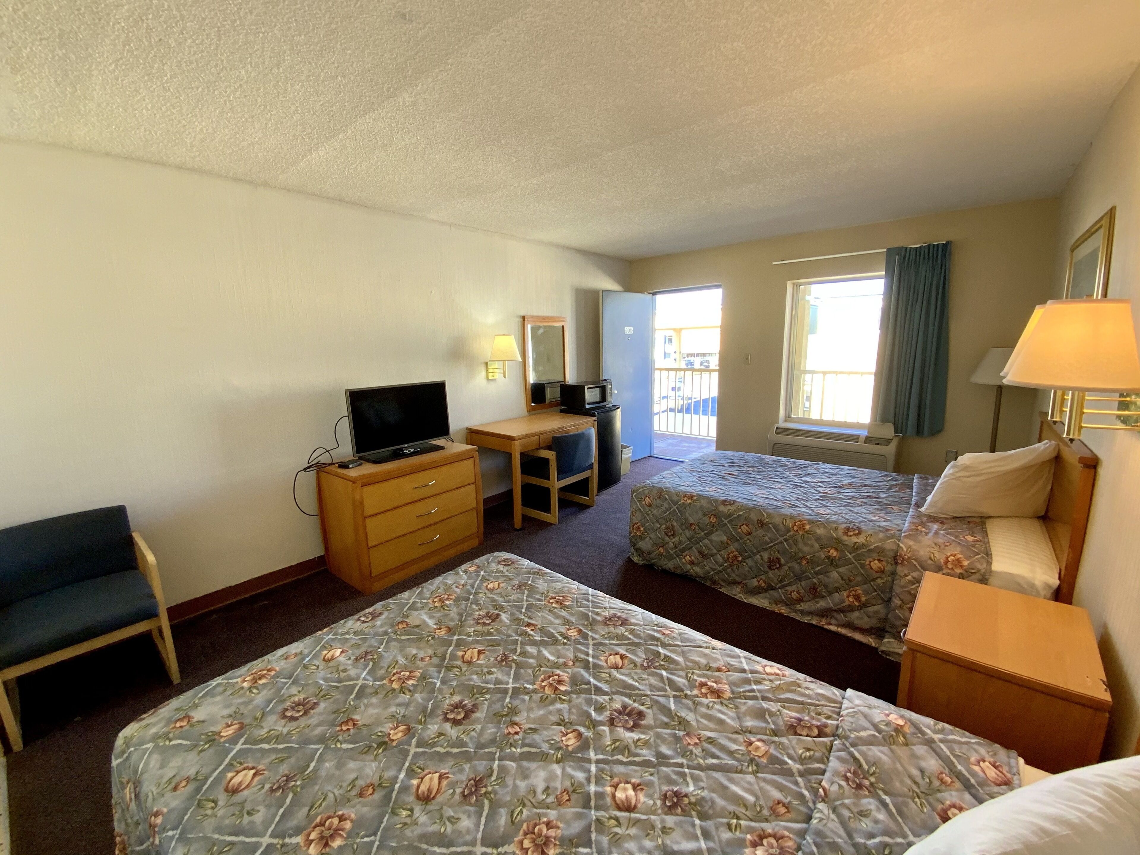 A hotel room with two beds, a desk, a television, and a small kitchenette.